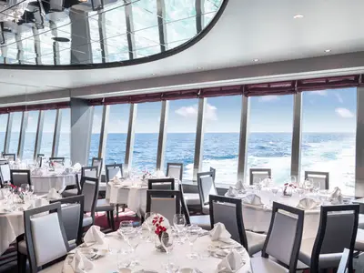 Das elegante Lighthouse Restaurant an Bord der MSC Bellissima zeigt mehrere Tische mit weissen Tischtuechern und Gedecken. Durch grosse Panoramafenster blickt man auf das weite, blaue Meer und den Horizont mit vereinzelten Wolken. An den Tischen stehen moderne Stühle bereit.