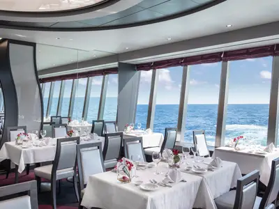 Elegantes Lighthouse Restaurant an Bord der MSC Bellissima. Hell gedeckte Tische mit Blumen vor großen Panoramafenstern mit weitem Blick auf das blaue Meer und den Himmel.