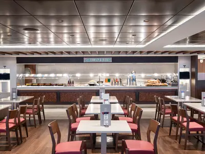 Blick in den hellen und modernen Buffetbereich 'Marketplace Buffet' an Bord der MSC Bellissima. Zahlreiche Tische und Stühle sind im Vordergrund zu sehen, während im Hintergrund eine lange Theke mit verschiedenen Speisen und Getränken bereitsteht, darunter ein 'Family & Kids'-Schild. Die Decke ist mit Metallpaneelen und eingelassenen Lichtern versehen.