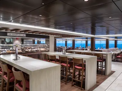 Ein modernes Buffetrestaurant, das Marketplace Buffet, an Bord der MSC Bellissima. Im Vordergrund lange Tische und Barhocker, dahinter eine Bäckerei-Auslage mit Gebäck. Große Fenster bieten einen weiten Blick auf das blaue Meer und den Horizont.
