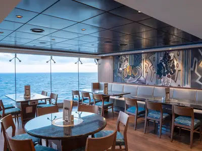 Geräumiges Restaurant an Bord der MSC Bellissima mit großen Panoramafenstern zum Meer. Es gibt Holztische mit blauen Stühlen und eine lange Sitzbank entlang einer Wand mit abstrakter Kunst.