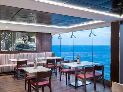 Modernes Buffet-Restaurant auf der MSC Bellissima. Helle Tische, rote Stühle und gestreifte Bänke. Große Fensterfront mit Meerblick. Abstraktes Kunstwerk an der Holzwand.