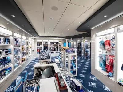 Blick in den modernen MSC Shop an Bord der MSC Bellissima. Beleuchtete Regale präsentieren MSC-Markenartikel wie Kleidung, Spielzeug und Souvenirs. Der blaue Teppich ist mit dem MSC Logo gemustert.