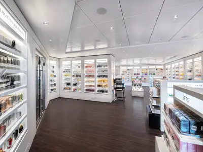 Ein heller, moderner Duty-free-Shop mit Parfüms und Kosmetika an Bord der MSC Bellissima. Beleuchtete Glasvitrinen zeigen verschiedene Markenprodukte wie Calvin Klein und Paco Rabanne. Der dunkle Holzboden und die weißen Wände schaffen eine elegante Atmosphäre. Keine Personen sichtbar.