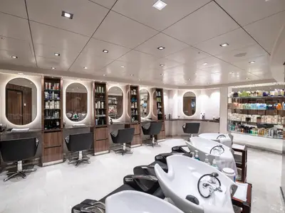 Moderner Friseursalon an Bord der MSC Bellissima. Styling-Stationen mit beleuchteten Spiegeln und Stühlen. Friseur-Waschbecken im Vordergrund. Wände mit Regalen und Pflegeprodukten.