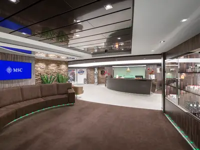 Blick in den Wartebereich und die Rezeption des MSC Aurea Spa an Bord der MSC Bellissima. Eine braune Rundcouch mit grüner LED-Beleuchtung im Vordergrund, davor ein großer Monitor mit dem MSC Logo. Im Hintergrund die Rezeption und Shops.