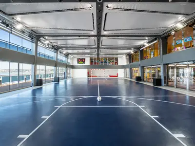 Ein weitläufiger, blauer Multifunktions-Sportplatz mit Basketball- und Fussballtoren an Bord der MSC Bellissima. Grosse Fenster bieten Panoramablick auf das Meer und eine Küstenlandschaft. Rechts befinden sich helle Lounges und Bars.