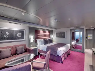 Moderne MSC Yacht Club Deluxe Suite an Bord der MSC Bellissima. Großes Doppelbett, Sofa und Spiegel, der den Blick auf den Balkon und das Meer reflektiert.