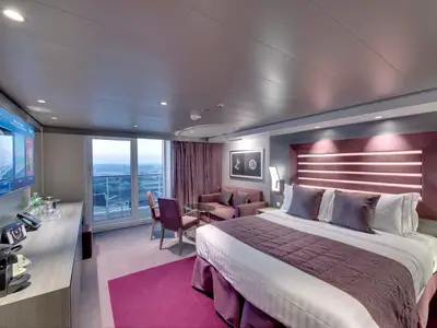 Eine luxuriöse MSC Yacht Club Deluxe Suite an Bord der MSC Bellissima. Das Zimmer zeigt ein großes Doppelbett, eine Sitzecke mit Sofa und Sesseln, sowie einen privaten Balkon mit Meerblick. Elegante Einrichtung in Magenta und Grau.