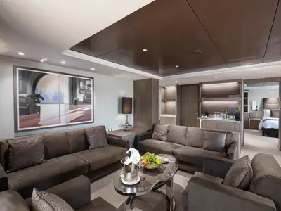Blick in den luxuriösen Wohnbereich einer Suite an Bord der MSC Bellissima. Zu sehen sind bequeme Sofas, ein Couchtisch mit Obst und Champagner, ein Beistelltisch mit Lampe und ein großes Kunstgemälde. Im Hintergrund ist ein angrenzendes Schlafzimmer sichtbar.