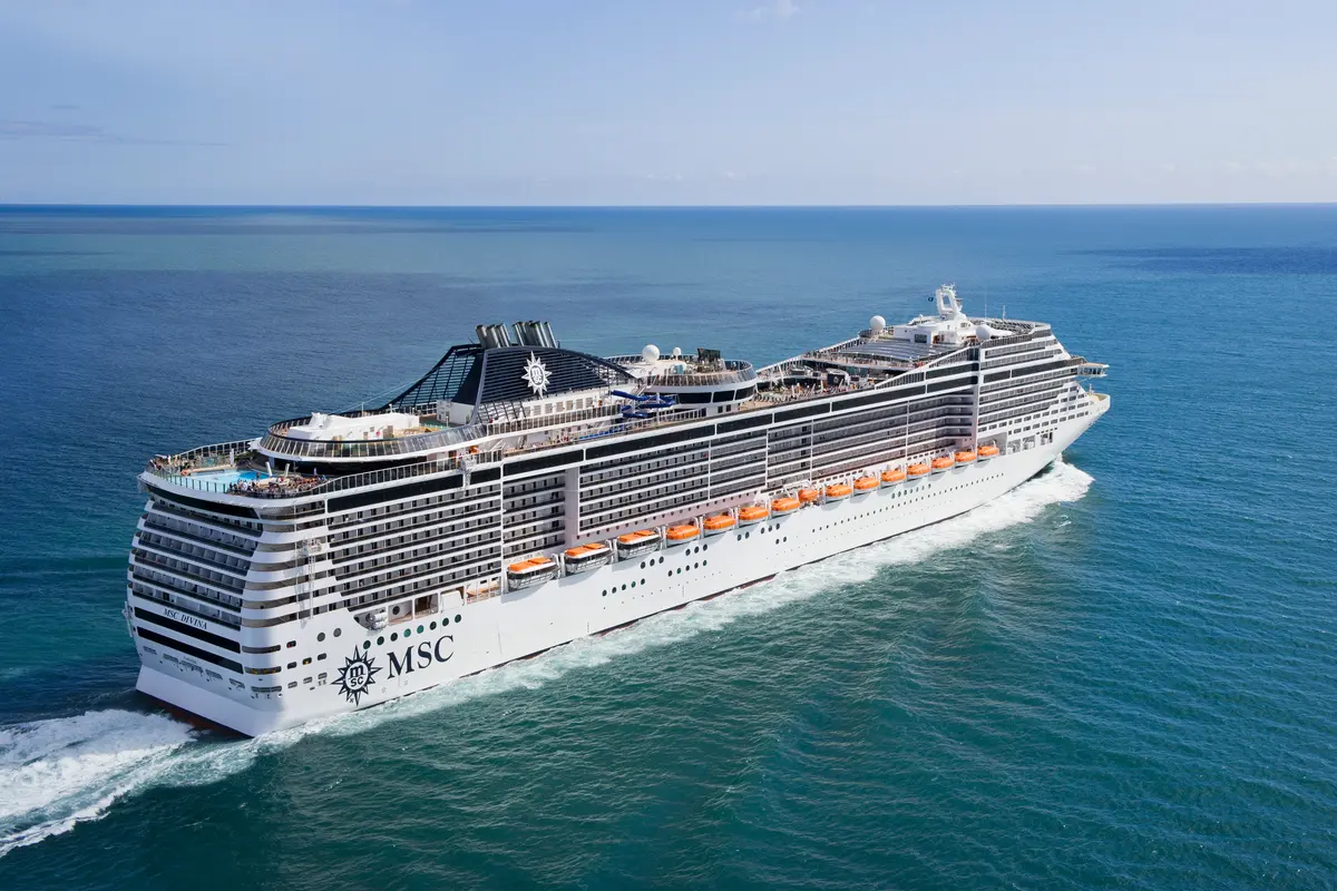 Die MSC Divina, ein grosses weisses Kreuzfahrtschiff, fährt auf dem tiefblauen Meer. Von oben gesehen, zeigt es die Decks, Pools und Schornsteine unter klarem, sonnigem Himmel.