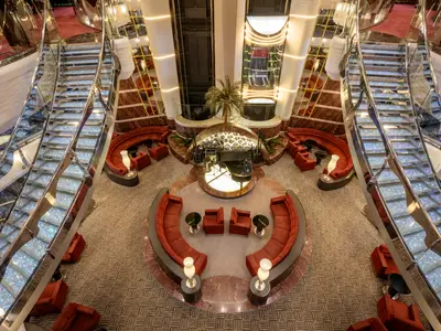 Blick von oben auf das luxuriöse Atrium der MSC Divina. Im Zentrum eine Pianobar, umgeben von roten Sitzgruppen. Zwei glitzernde Treppen führen nach oben.