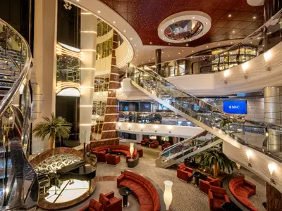 Einblick in das luxuriöse Atrium der MSC Divina. Geschwungene Treppen, ein Piano, rote Sofas und Palmen schaffen eine elegante Atmosphäre. MSC-Logo auf dem Bildschirm.
