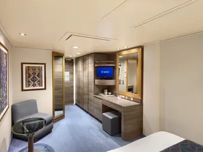 Helle Aussenkabine der MSC Divina mit blauem Teppich. Eine Sitzecke mit Sessel und Glastisch ist links zu sehen. Rechts befinden sich ein Schreibtisch mit TV, Spiegel und Hocker sowie Schränke für Stauraum.