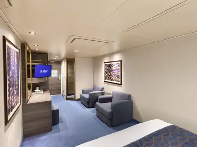 Helle Aussenkabine der MSC Divina. Komfortabler Sitzbereich mit zwei blauen Sesseln, Waschtisch und TV. Moderne Kunstwerke an den Wänden, blauer Teppichboden. Einladendes Ambiente.