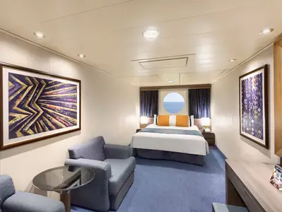 Eine elegant eingerichtete Aussenkabine an Bord der MSC Divina mit grossem Doppelbett, zwei Sesseln und einem runden Bullauge mit Blick auf das Meer. Violett-goldene Kunstwerke schmuecken die Waende.