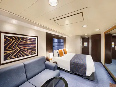Ansicht einer modernen, eleganten Kabine an Bord der MSC Divina mit großem Doppelbett, blauer Tagesdecke, orangen Kissen, einem blauen Sofa und einem abstrakten Wandbild.