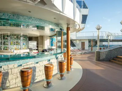 Sonniges Deck mit der Poseidon Bar an Bord der MSC Divina. Die halbrunde Bar hat eine gläserne Theke, blaue Fliesen und Holzbarhocker. Im Hintergrund Liegestühle und der offene Meerblick.