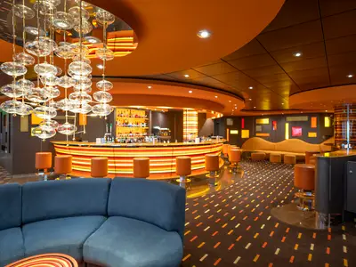 Die elegante Golden Jazz Bar an Bord der MSC Divina zeigt eine geschwungene Bar mit orangefarbenen Hockern. Ein großer gläserner Kronleuchter beleuchtet den Raum. Das Interieur in warmen Orange- und Brauntönen umfasst auch gemütliche Sofas und einen gemusterten Teppich.