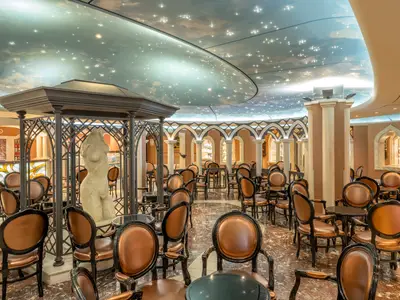 Die elegant gestaltete Piazza Del Doge Bar an Bord der MSC Divina. Der Raum ist mit vielen runden Tischen und braunen Ledersesseln ausgestattet. Die Decke zeigt einen Sternenhimmel und Wolken. Im Zentrum steht eine Skulptur in einem Metallpavillon.