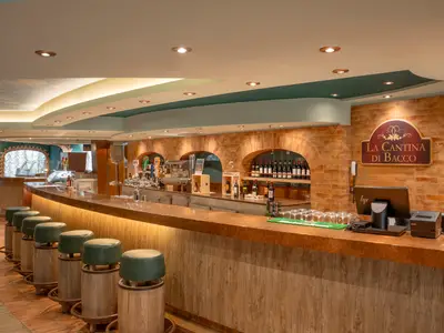 Die rustikale Bar 'La Cantina di Bacco' an Bord der MSC Divina. Ein langer Tresen mit Holz-Barhockern erstreckt sich durch den Raum. Im Hintergrund sind Ziegelsteinbögen mit Weinregalen, Bierhähnen und einer Kasse zu sehen. Warmes Licht betont das gemütliche Ambiente und die gewölbte Decke.