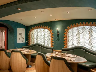 Einladender Blick in das Restaurant La Cantina di Bacco an Bord der MSC Divina mit mehreren Tischen, grünen Lederbänken und Holzsesseln. Die Wände sind türkisgrün, akzentuiert durch Fenster in Ziegelsteinbögen und eine Weinkarte an der Wand.