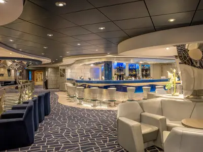 Die Bar an Bord der MSC Divina zeigt geschwungenen Tresen, Barhocker, beleuchtete Alkoholregale, elegante Sofas, Sessel und blauen Musterteppich.