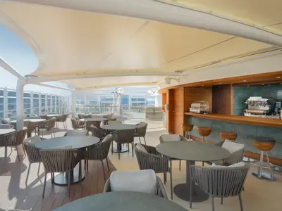 Einladende The One Bar an Bord der MSC Divina mit zahlreichen Tischen, Rattan-Stuehlen und Bartresen unter einem hellen Sonnensegel. Im Hintergrund ist das Meer sichtbar.
