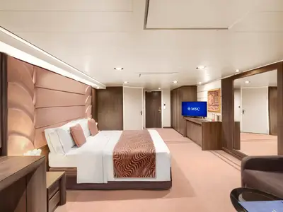 Eine helle und geräumige Suite an Bord der MSC Divina. Ein großes Doppelbett mit rosafarbenen Kissen und einer gemusterten Tagesdecke dominiert den Raum. Rechts steht ein Flachbildfernseher und ein dunkles Sideboard. Links ein kleiner Schminktisch. Die Kabine ist modern und elegant eingerichtet.