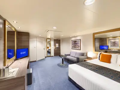 Geräumige barrierefreie Suite an Bord der MSC Divina mit Doppelbett, Sofa, TV und Spiegel. Helle Einrichtung mit Holzpaneelen, blauem Teppich und orangen Kissen.