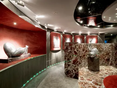 Elegante Kunstgalerie an Bord der MSC Divina. Moderne Skulpturen, rote Wände, Marmorboden und grüne LED-Beleuchtung definieren das luxuriöse Design.