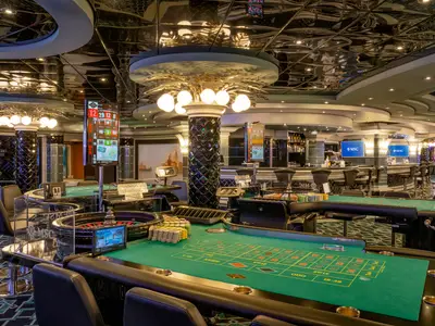 Casinò Veneziano auf der MSC Divina. Grüne Spieltische, Rouletterad, Chips vorne. Hinten Spielautomaten und Bar. Glänzende Decke reflektiert elegantes Licht.