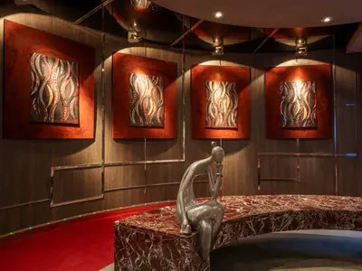 Die elegante Kunstgalerie an Bord der MSC Divina. Eine silberne Skulptur sitzt auf einem roten Marmortisch. Vier abstrakte, beleuchtete Kunstwerke in Rot und Schwarz schmücken die holzvertäfelten Wände. Roter Teppichboden.