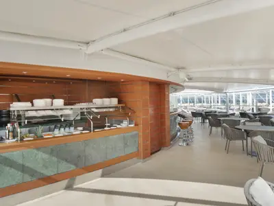 Ein heller, überdachter Buffet- und Barbereich an Bord der MSC Divina mit Holzelementen, einer Essensauslage und zahlreichen Sitzgelegenheiten im Freien. Im Hintergrund ist das Meer sichtbar.