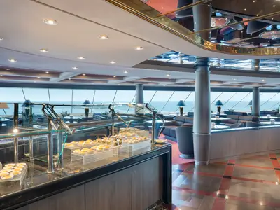 Ein eleganter Lounge-Bereich mit einem reichhaltigen Buffet an kleinen Speisen und Desserts an Bord der MSC Divina. Große Fenster bieten einen weiten Blick auf das Meer.