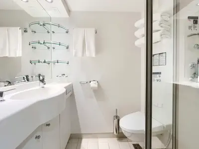 Ein modernes Badezimmer an Bord der MSC Divina. Der Waschtisch mit Seifenspender und Armatur ist links, die Toilette und Duschkabine rechts. Weiße Handtücher hängen an der Wand und liegen gestapelt im Duschbereich. Helle, minimalistische Einrichtung.