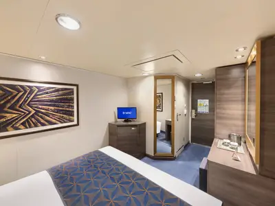 Blick in eine moderne Innenkabine an Bord der MSC Divina. Ein Doppelbett mit blauem Überwurf, TV, Ganzkörperspiegel und ein kleiner Schreibtisch sind zu sehen.