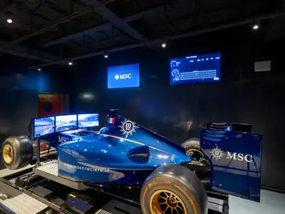 Ein leuchtend blauer Formel-1-Simulator, der MSC Formula Racer, an Bord der MSC Divina. Er steht in einem dunklen Raum mit Bildschirmen, die eine Rennstrecke und Live-Zeiten zeigen. Goldene Felgen sind sichtbar.