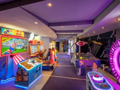 Innenansicht der Virtual World Arcade an Bord der MSC Divina. Bunte Spielautomaten, darunter ein Schiessspiel und Airhockey, beleuchten den Gang mit Neonlicht. Der Raum ist menschenleer.