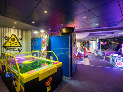 Ein farbenfroher Spielbereich mit Airhockeytischen und anderen Arcade-Automaten an Bord der MSC Divina, beleuchtet von Neonlichtern. Eine Wand zeigt ein 'Danger'-Graffiti mit Totenkopf.
