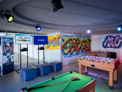 Der helle Teenager-Club an Bord der MSC Divina mit Billardtisch, zwei Kickertischen, blauen Sitzwürfeln und großen Bildschirmen. Die Wände sind mit farbenfrohen Graffiti bemalt und unterstreichen die jugendliche Atmosphäre.