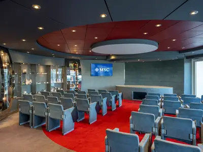 Konferenzraum der MSC Divina. Graue Stühle auf rotem Teppich, großer Bildschirm mit MSC Logo. Seitliche Produkt-Vitrinen. Modernes Design mit dunkler Decke und roten Akzenten.