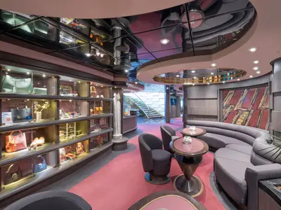 Blick in eine elegante Lounge an Bord der MSC Divina. Links eine Vitrinenwand mit Handtaschen und Accessoires. Rechts geschwungene Sofas und Tische, im Hintergrund eine Treppe.
