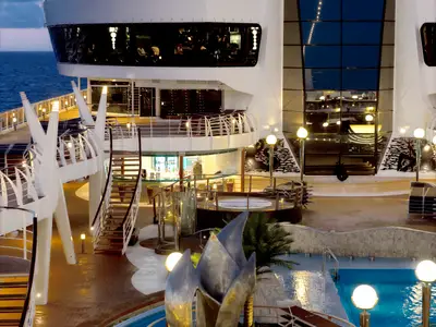 Abendlicher Aussenbereich der MSC Divina. Beleuchteter Pool, Whirlpool, Bars und elegante Treppen auf mehreren Decks. Moderne Architektur und Lichtinstallationen bei Nacht.