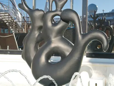 Eine dunkle, abstrakte Skulptur mit weichen Formen und Löchern ist auf dem Sonnendeck der MSC Divina zu sehen. Im Hintergrund spiegeln sich Schiffstrukturen wider.