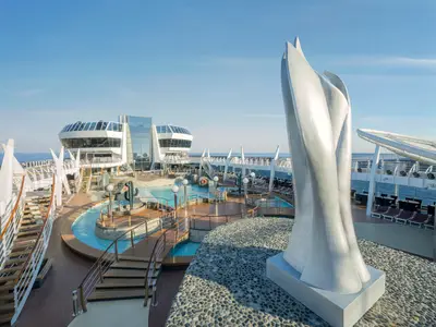 Das sonnige Sonnendeck der MSC Divina mit Aquapark, Pools und Lounge-Bereichen. Eine grosse, moderne Skulptur prägt die Szene. Im Hintergrund: weites Meer unter blauem Himmel. Keine Personen sind sichtbar.
