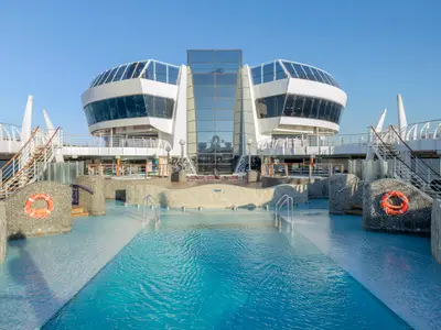Der Aquapark der MSC Divina zeigt Pools und futuristische Glasarchitektur auf den Oberdecks, umgeben von Liegeflächen und Treppen unter blauem Himmel. Keine Personen sichtbar.