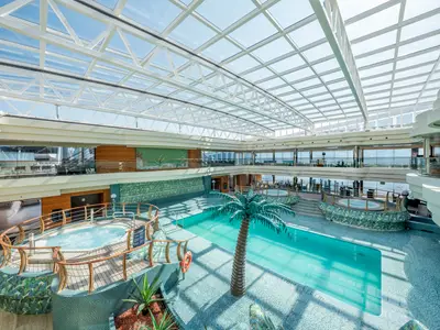 Panoramablick auf den überdachten Poolbereich „Le Sirene“ der MSC Divina mit großem Pool, Whirlpools, Palmen und Glasdach.