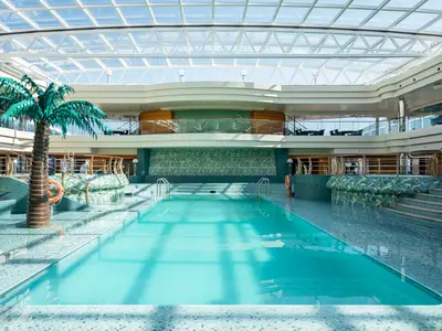 Blick auf den grossen Innenpool Le Sirene an Bord der MSC Divina. Der Poolbereich ist von einem gläsernen Dach bedeckt und mit grünen Kacheln und einer Palmen-Skulptur tropisch gestaltet.