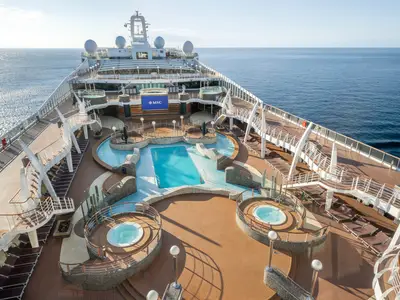 Blick von oben auf den Aquapark mit Pools, Whirlpools und Sonnenliegen an Bord der MSC Divina bei Sonnenschein. Das Meer ist im Hintergrund sichtbar.